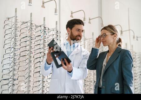 Optometristo maschio che aiuta la donna a scegliere gli occhiali nel negozio ottico che mostra al suo specchio Foto Stock