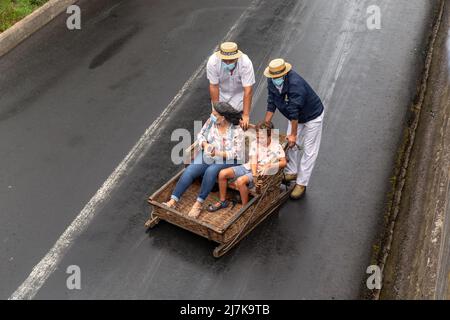 FUNCHAL, PORTOGALLO - 24 AGOSTO 2021: Donna e bambino non identificati rotolare giù per la strada in cesti di vimini (toboga) con l'aiuto dei conducenti. Foto Stock