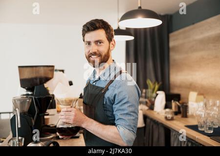 Sorridente barista che tiene in mano il bollitore con caffè preparato e guarda la fotocamera Foto Stock