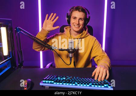 Il giovane e bel giocatore professionista vlogger che si allena mano alla fotocamera, dice ciao ai suoi iscritti e seguaci, sorridendo con la fotocamera. Registra in diretta streaming di videogiochi a casa di notte. Cyber sport Foto Stock