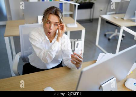 Donna da ufficio che regola il mascara davanti agli occhi mentre guarda la fotocamera dello smartphone Foto Stock