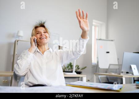 Donna d'affari che parla al telefono e agita le mani per salutare i suoi colleghi Foto Stock