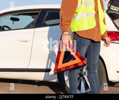 uomo irriconoscibile sul gilet che tiene un triangolo rosso sulla strada con l'automobile rotta e concetto di guasto Foto Stock