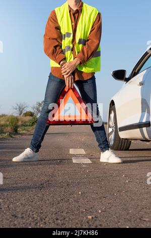 Vista frontale verticale di giovani uomini irriconoscibili che tengono un triangolo rosso d'allarme al centro delle gambe aperte, con un'auto bianca rotta sul lato Foto Stock