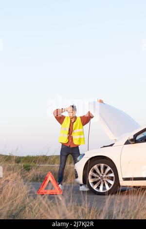 Foto verticale di giovani uomini in gilet che cercano aiuto sulla strada con la sua auto rotta di fronte a lui nel pomeriggio Foto Stock