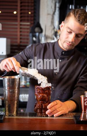 Concentrato giovane barista maschile alla moda in uniforme versare cubetti di ghiaccio in tiki bicchiere mentre si prepara un cocktail rinfrescante al banco del bar Foto Stock