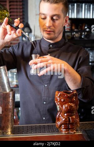 Giovane barman maschile con acconciatura alla moda in uniforme versando ron in jigger mentre si trova al banco e preparare cocktail alcolici al bar Foto Stock