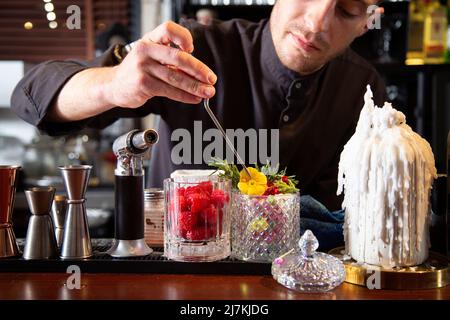 Serio barkeeper in uniforme nera che decora cocktail con fiori freschi durante il lavoro al bar Foto Stock