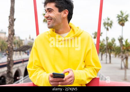 Giovane uomo allegro con cappuccio giallo che sorride mentre tiene il telefono mentre riposa all'aperto in strada Foto Stock
