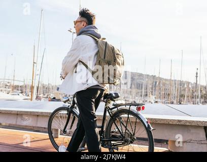 Vista laterale del maschio con zaino passeggiando in bicicletta nel porto vicino al mare con navi ormeggiate in giornata di sole nella città costiera Foto Stock