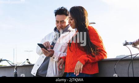 Coppia multirazziale in abbigliamento casual surf internet su cellulare mentre si trova vicino recinzione e biciclette su strada soleggiata della città Foto Stock