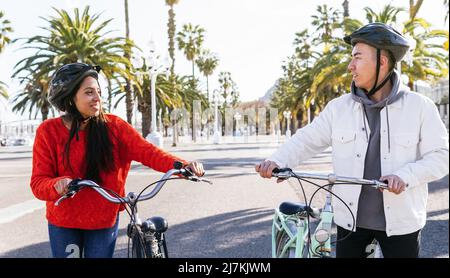 Coppia felice in caschi a piedi con biciclette guardarsi l'un l'altro su strada asfaltata in città con tress verde il giorno d'estate Foto Stock