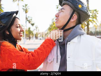 Vista laterale del positivo Indian girlfriend che fissa casco protettivo sul ragazzo asiatico mentre si alza insieme su strada con alberi verdi Foto Stock