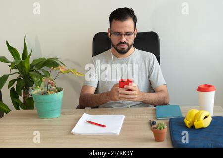 L'uomo si è concentrato nella messaggistica di testo degli auricolari sullo smartphone e nel lavoro di documentazione durante il lavoro a tavola in ufficio Foto Stock