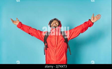 Felice uomo turistico bearded in giacca rossa e zaino con le braccia sollevate su sfondo blu in studio chiaro Foto Stock