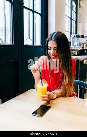 Bella sorridente donna moderna godendo di succo di frutta mentre si siede al tavolo di legno in un caffè Foto Stock