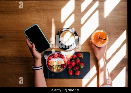 Vista dall'alto del raccolto femminile con smartphone e coppa di contenimento di succo d'arancia mentre si siede a tavola di legno e avere deliziosa colazione sana con fru Foto Stock