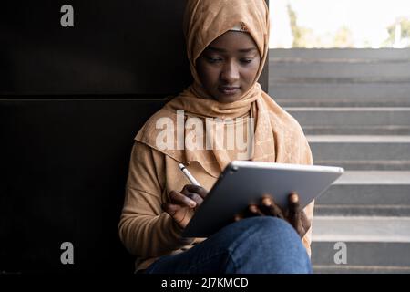 Concentrato giovane studentessa musulmana afroamericana in foulard tradizionale seduta sulla strada che naviga su tablet con stilo mentre prepara fo Foto Stock