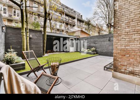 Sedie su cortile piastrellato di mattoni edificio residenziale situato vicino prato erboso con garage contro l'alloggio in città il giorno d'estate Foto Stock