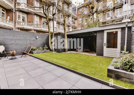 Sedie su cortile piastrellato di mattoni edificio residenziale situato vicino prato erboso con garage contro l'alloggio in città il giorno d'estate Foto Stock