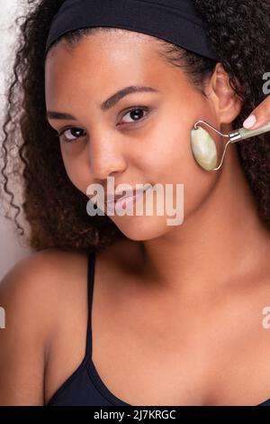 Contenuto giovane etnia femminile con pelle perfetta e capelli ricci lunghi e scuri fascia guardando la macchina fotografica mentre si fa il massaggio viso con jade rulli di nuovo Foto Stock