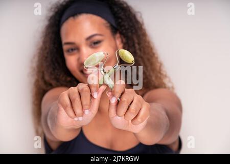 Felice giovane ispanica femminile millennial con capelli ricci lunghi e fascia sorridente e dimostrare verde viso jade rulli in studio luce Foto Stock