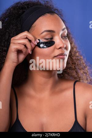 Giovane ispanica femmina modello con lunghi capelli ricci scuri una fascia sorridente felicemente e guardando via mentre si applicano le macchie occhi su sfondo blu Foto Stock