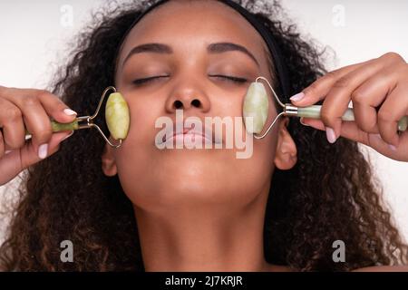 Contenuto giovane etnia femminile con pelle perfetta e capelli ricci lunghi e scuri che chiudono gli occhi mentre si fa massaggio viso con jade rulli contro bianco backgroun Foto Stock