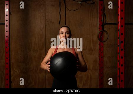 Sportivo concentrato giovane fit in abbigliamento sportivo in piedi con palla di medicina pesante e guardando la macchina fotografica durante l'allenamento in palestra Foto Stock