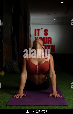 Donna giovane e aggraziata in abbigliamento sportivo che si esibisce in posa Bhujangasana mentre pratica yoga sul tappeto in palestra moderna Foto Stock