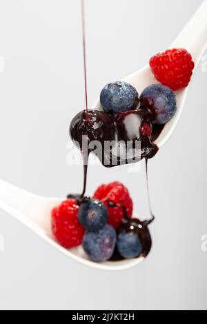 Condimento dolce al cioccolato versando su un mucchio di lamponi freschi e maturi mirtilli su cucchiai su sfondo bianco in studio chiaro Foto Stock