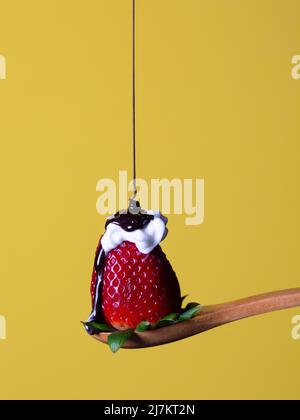 Sciroppo di cioccolato dolce versato su fragola rossa matura con panna bianca montata su cucchiaio di legno su sfondo giallo in studio chiaro Foto Stock