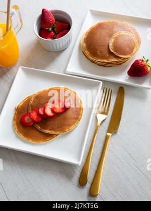 Vista dall'alto delle frittelle fresche servite su piatti bianchi con fragole mature su sfondo grigio con succo d'arancia in cucina Foto Stock