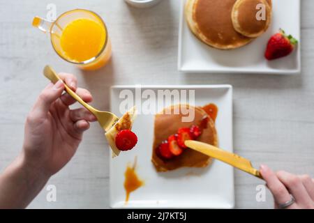 Vista dall'alto di raccolto anonima persona taglio gustosi pancake dolci con fragole servito su sfondo grigio con succo d'arancia in cucina Foto Stock