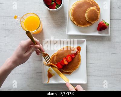 Vista dall'alto di raccolto anonima persona taglio gustosi pancake dolci con fragole servito su sfondo grigio con succo d'arancia in cucina Foto Stock