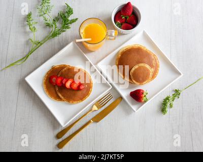 Vista dall'alto delle frittelle fresche servite su piatti bianchi con fragole mature su sfondo grigio con succo d'arancia in cucina Foto Stock