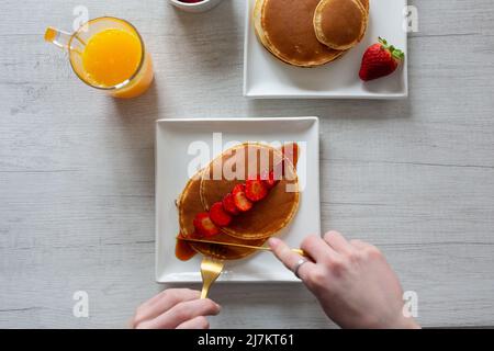 Vista dall'alto di raccolto anonima persona taglio gustosi pancake dolci con fragole servito su sfondo grigio con succo d'arancia in cucina Foto Stock