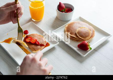 Da sopra di raccolto anonima persona taglio gustosi pancake dolci con fragole servita su sfondo grigio con succo d'arancia in cucina Foto Stock