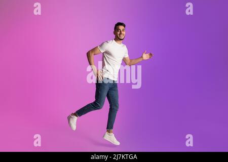 L'intera lunghezza del giovane arabo felice in t-shirt e jeans che corrono in luce al neon, spazio copia Foto Stock