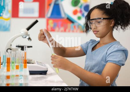 Studentessa elementare biraciale che indossa occhiali protettivi facendo esperimenti chimici in laboratorio. Inalterata, educazione, apprendimento, sperimentazione scientifica Foto Stock