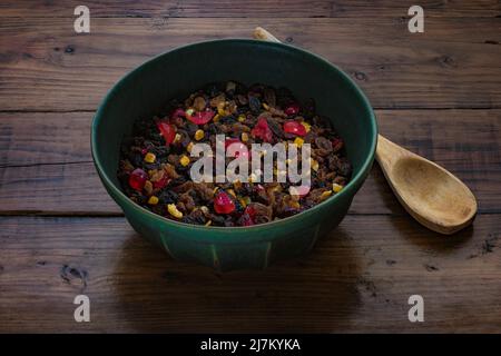 Recipiente di miscelazione verde scuro contenente frutta secca mista, insieme a un cucchiaio di legno su una superficie di lavoro di legno scuro. Foto Stock