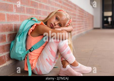 Ritratto di scolaretta elementare caucasica con zaino seduto sul pavimento nel corridoio della scuola Foto Stock