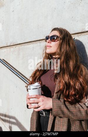 Donna elegante in outerwear con occhiali da sole appoggiati a parete e guardando via mentre si gusta il caffè per andare e sognare sulla strada della città Foto Stock