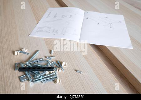 Parti di mobili non finiti, viti metalliche e utensili giacenti sul pavimento con manuale di istruzioni per l'assemblaggio di mobili sullo sfondo. Passare a. Foto Stock