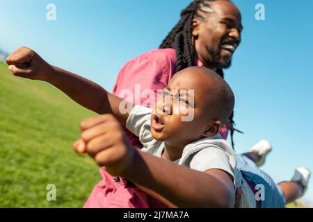 Il padre africano americano che porta il figlio mentre il ragazzo imita come supereroe che vola in giorno di sole Foto Stock