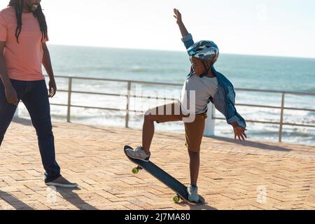 Ragazzo afroamericano che mostra abilità skateboard a padre sul lungomare durante la giornata di sole Foto Stock