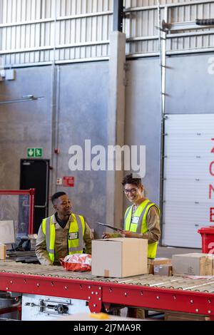 Sorridente giovane americano africano e collaboratore maschio asiatico maturo che controlla i pacchi sul nastro trasportatore Foto Stock