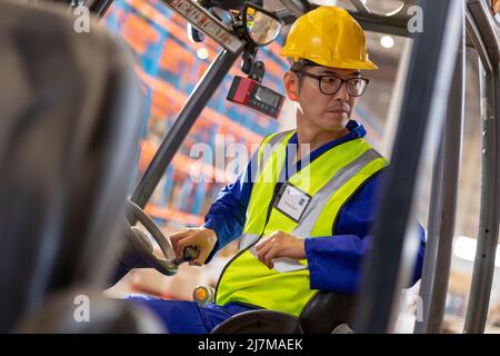 Serio lavoratore asiatico maturo di sesso maschile guardando via mentre si siede in carrello elevatore a magazzino Foto Stock