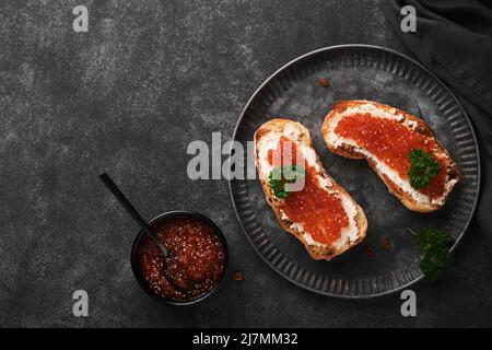 Due dandwiches con caviale rosso. Caviale rosso salmone in ciotola e sandwich server su vecchio piatto di ferro su sfondo vecchio tavolo nero. Vista dall'alto. Spazio di copia. Foto Stock