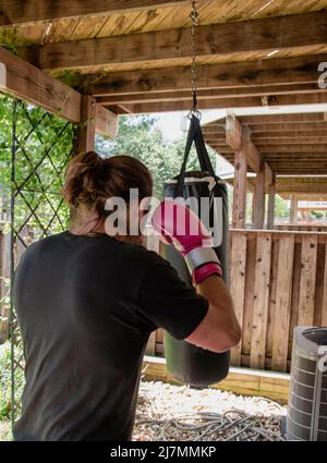 Un uomo che si esercita in una palestra domestica esterna, usando i guanti di boxe rosa luminosi. Borsa da punzonatura appesa al balcone sopra. Foto Stock